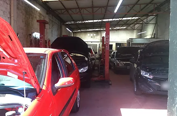 Carros com o capô aberto lado a lado nos boxes da oficina, sob a estrutura metálica do galpão.