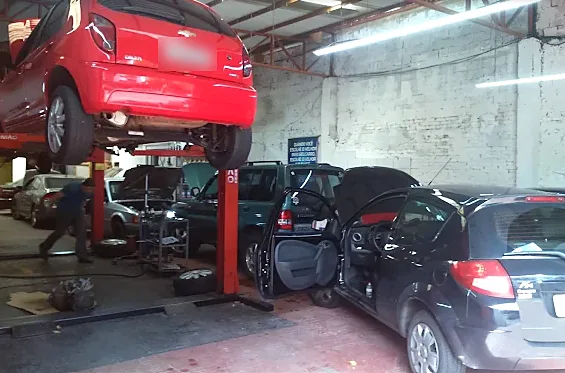 Interior da oficina: carro vermelho erguido no elevador e outros veículos em atendimento no pátio coberto.